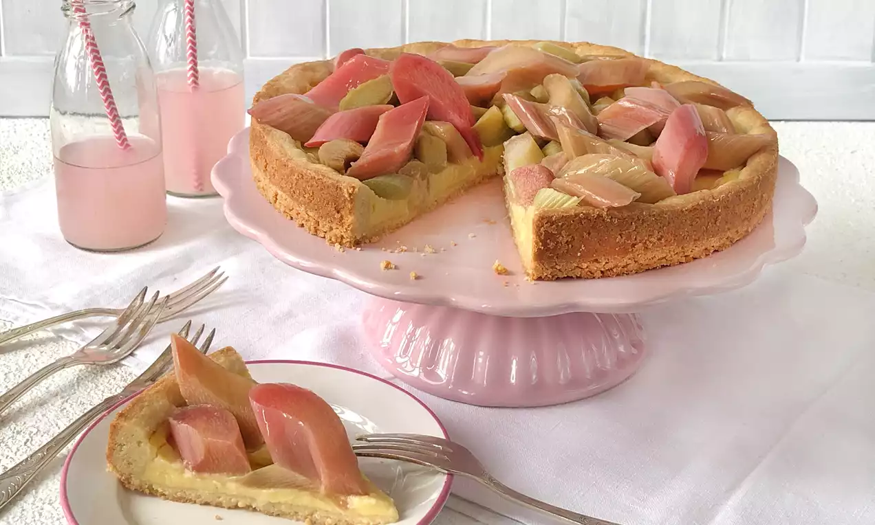 Cremige Rhabarbertarte mit goldbraunem Mürbeteigboden, rosaroten Rhabarberstücken und heller Füllung auf rosa Tortenplatte mit angeschnittenem Stück