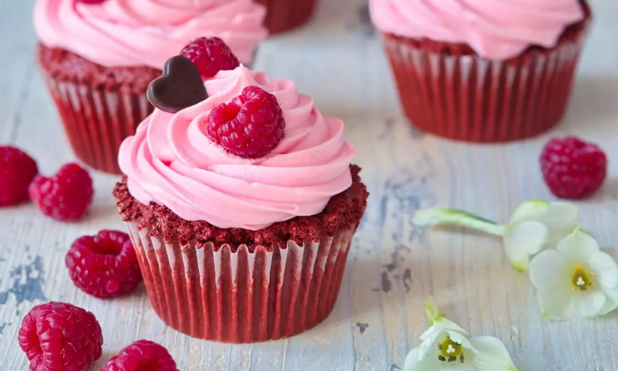 Cupcakes s červeným korpusom a ružovým krémom do špirály, ozdobené malinou a čokoládovým srdiečkom, na drevenom podklade s kvetmi