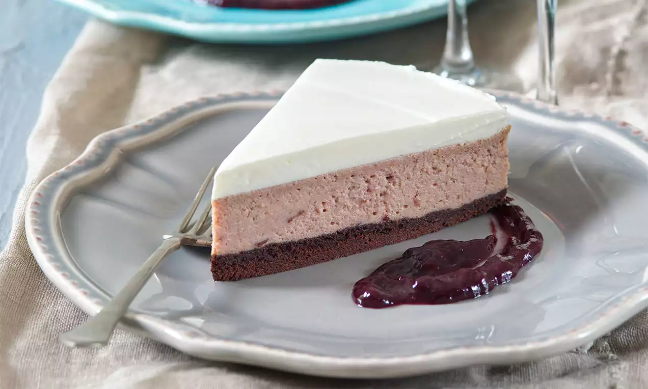 Trovrstvový koláč s tmavým kakaovým základom, jemnou hnedou plnkou, krémovo bielym vrchom a lyžicou lesklých povidiel na okrajovom tanieri