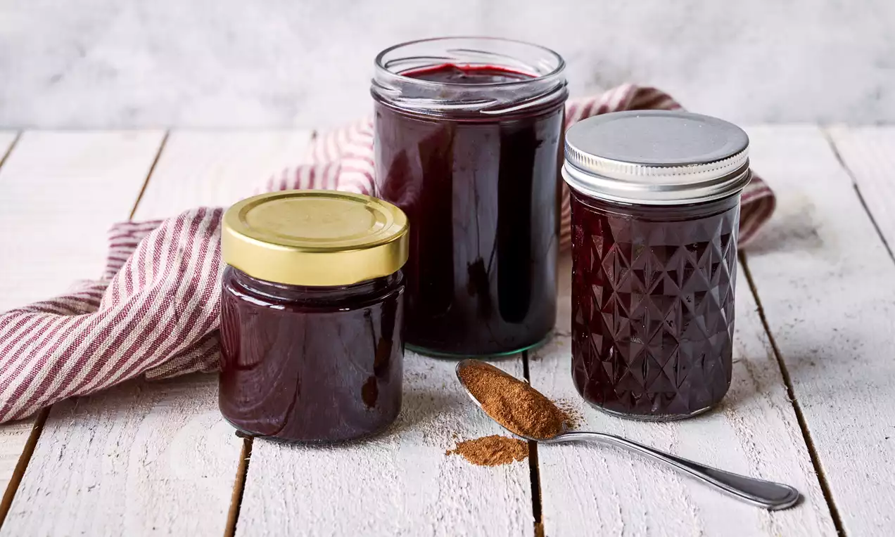 Tre glas med mørkerød julemarmelade placeret på lyst træbord med rødstribet klæde og en sølvske med brun krydderi ved siden af