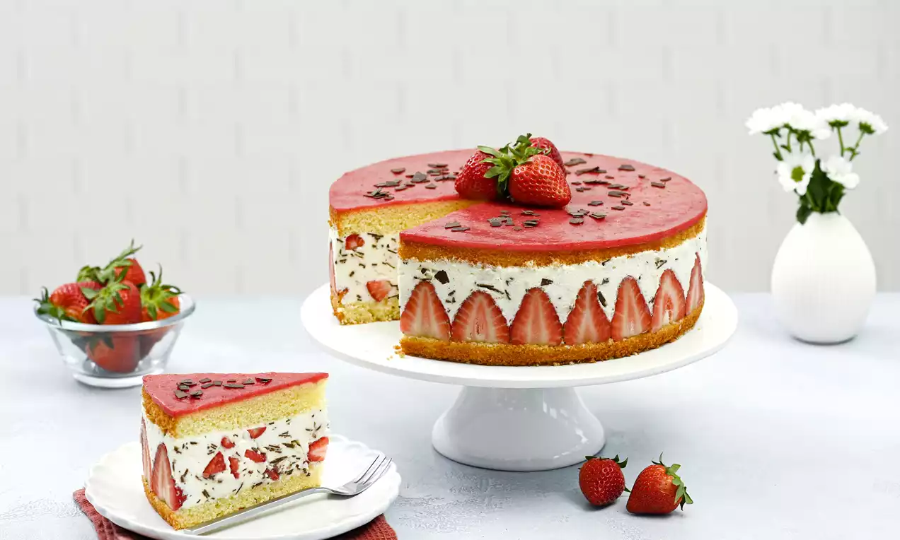 Erdbeer-Stracciatella-Torte mit roter Fruchtspiegelschicht, eingerahmten Erdbeeren und Schokosplittern auf weißer Tortenplatte angerichtet
