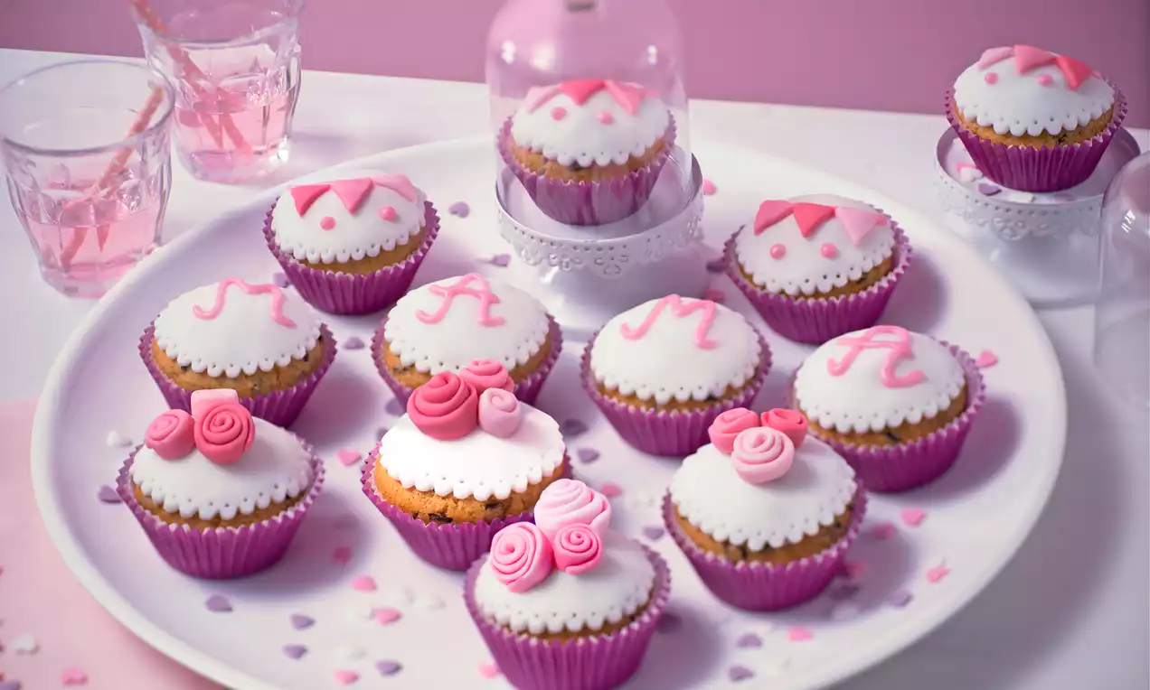Rosa Muffins mit weißem Zuckerguss, dekoriert mit Buchstaben und Zuckerröschen, in pinken Förmchen auf weißer Platte angerichtet