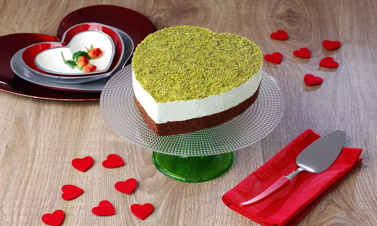 Kalp şeklinde cam ayaklı servis tabağında yer alan ve üstü ince öğütülmüş antep fıstığıyla kaplı, beyaz kremalı pasta