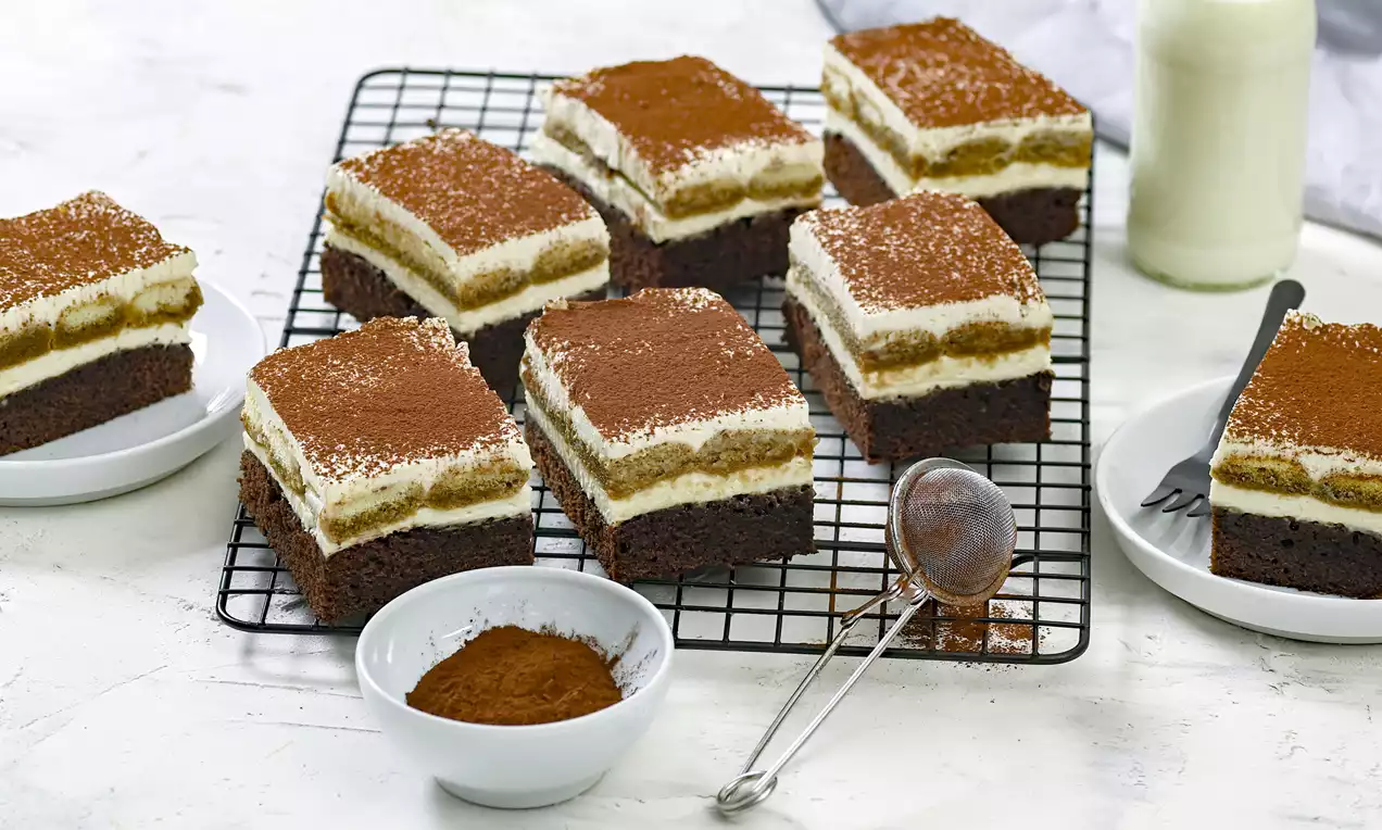 Quadrados com base de brownie escuro, creme claro, camada de café polvilhada com cacau em grade metálica sobre superfície branca