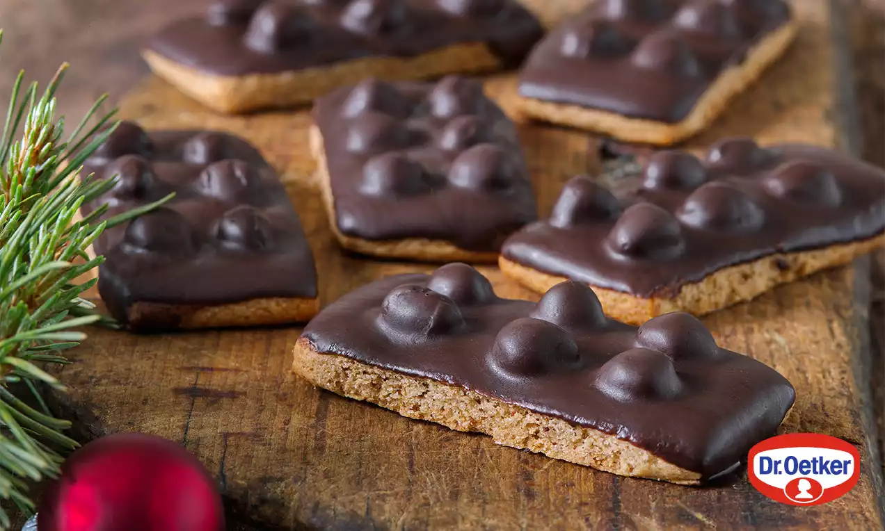 Hnedé obdĺžnikové sušienky potiahnuté hladkou tmavou čokoládou s oblými výstupkami, uložené na drevenom povrchu vedľa vetvičky jedle