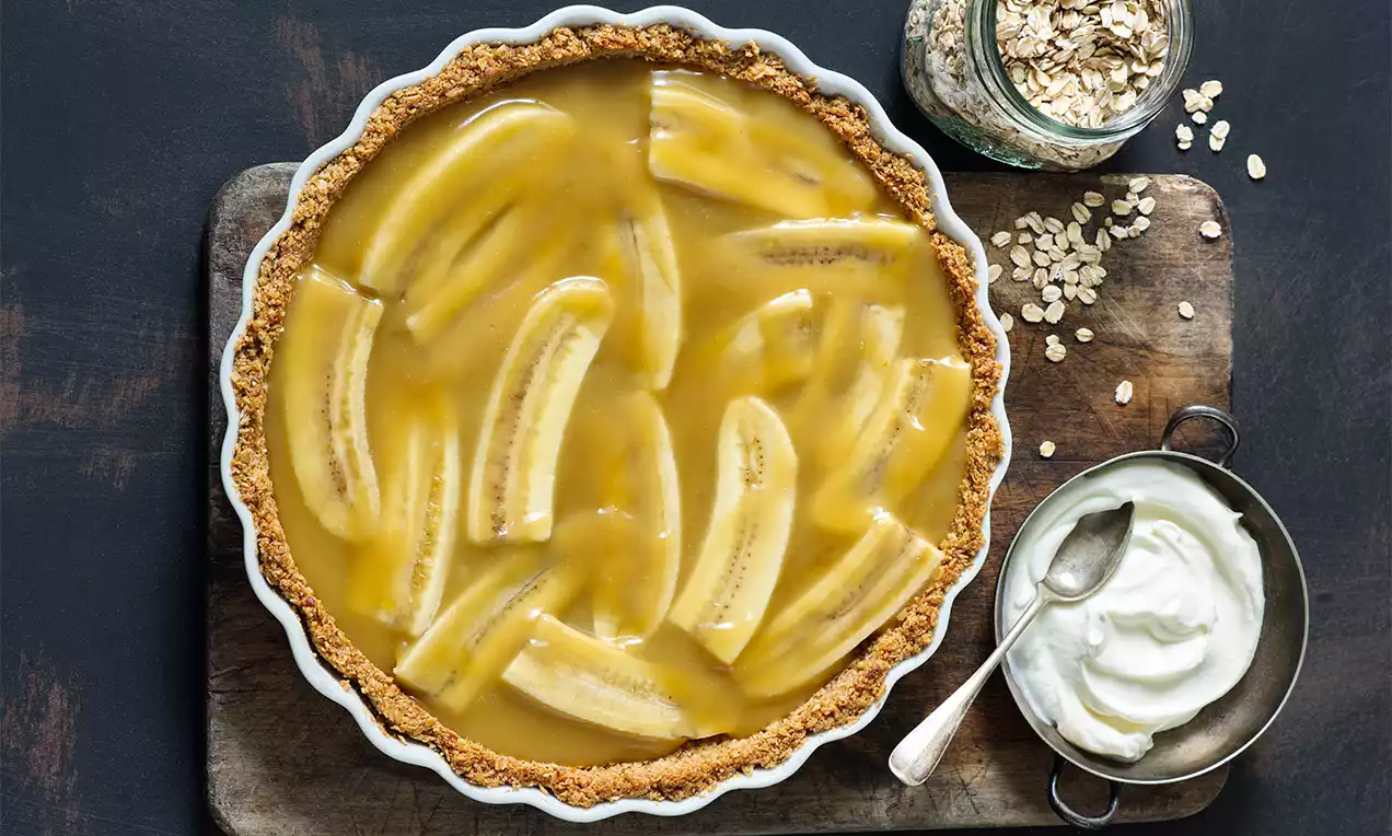 Vegan Banoffee s křupavým hnědozlatým korpusem, plátky banánů zalité karamelovou polevou, servírovaný na tmavém podnose s miskou šlehačky