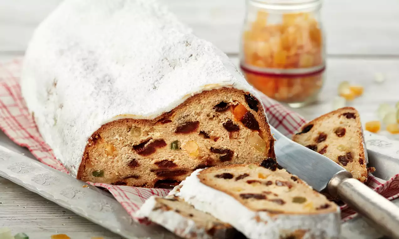 Butterstollen mit heller, bemehlter Kruste, lockerer Struktur und sichtbar eingebackenen Früchten auf einem Holztablett mit rotem Tuch drapiert