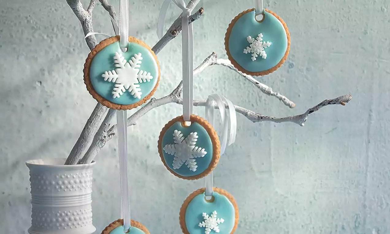 Runde Schneeflocken-Kekse mit hellblauem Zuckerguss und weißen Schneeflocken, dekorativ an weißen Zweigen mit Bändern aufgehängt