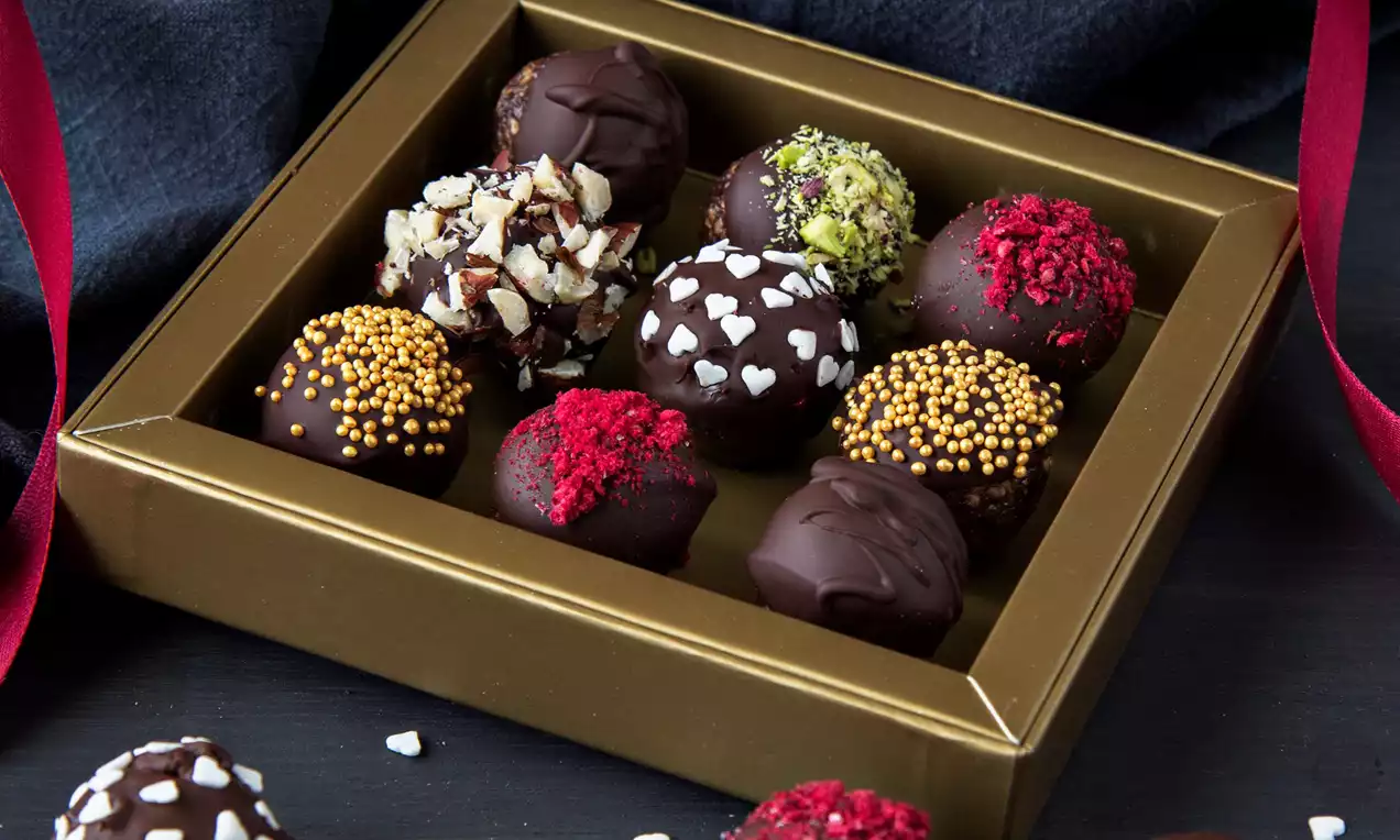 Tmavohnedé čokoládové pralinky zdobené farebnými posýpkami, srdiečkami a orieškami, uložené v zlatej krabičke na čiernom podklade.
