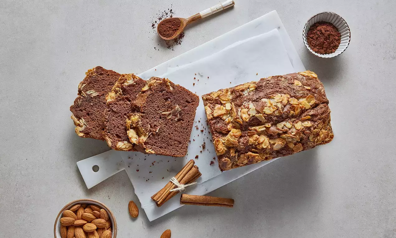 Kakao rengi nemli kek, altın rengi badem parçacıkları ve kıvrımlı tarçın deseni ile mermer görünümlü beyaz bir tabakta dilimlenmiş