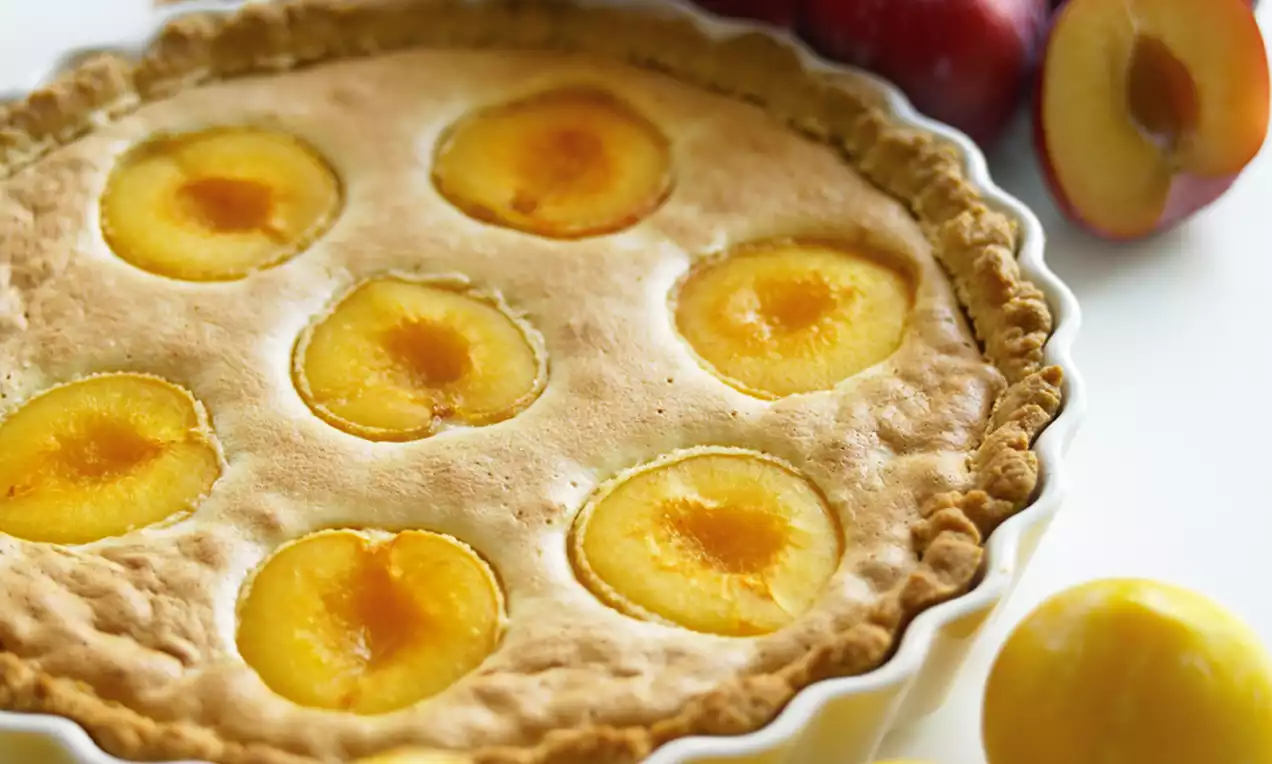 Luumupiiras vaaleassa keramiikkavuoassa, kullankeltaista täytettä ruskean rapean reunan ympäröimänä, taustalla kokonaisia ja puolikkaita luumuja