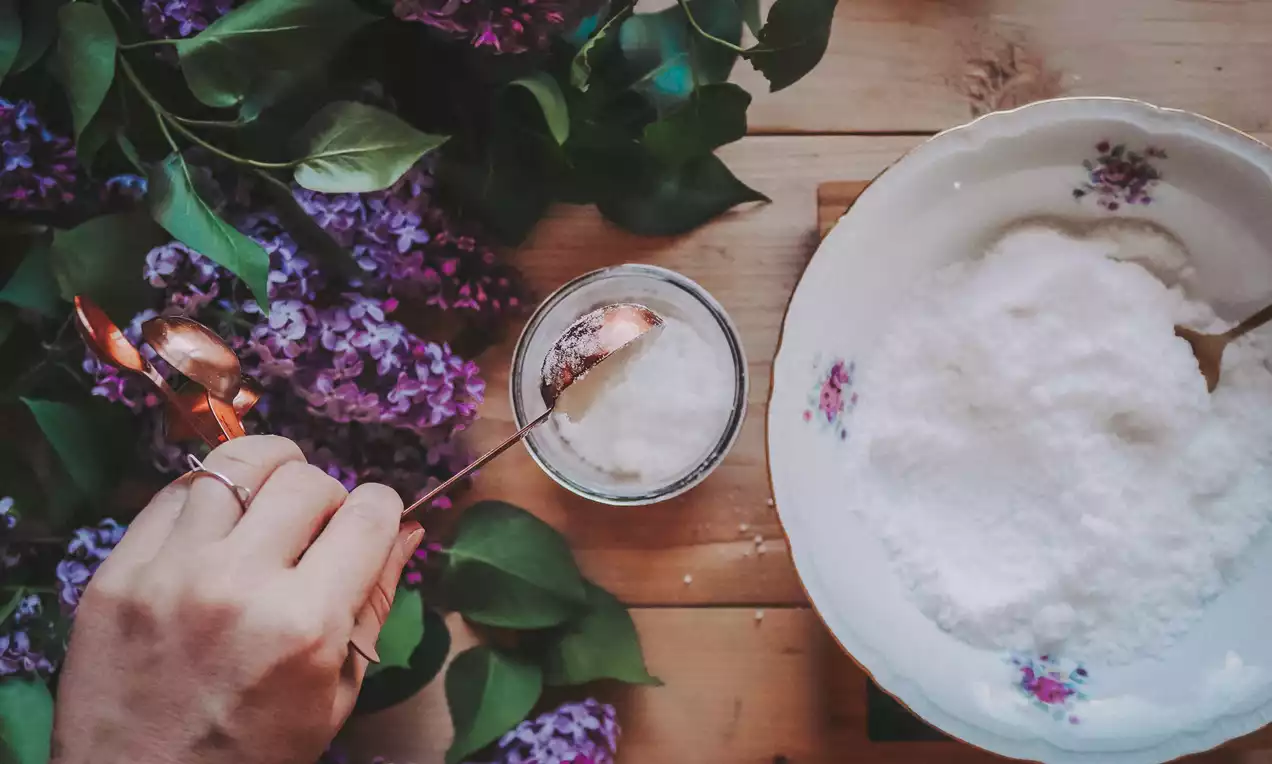 Bílý jemný prášek v porcelánové misce s květinovým motivem, vedle sklenička a bronzová lžička, okolí zdobí fialové květy šeříku