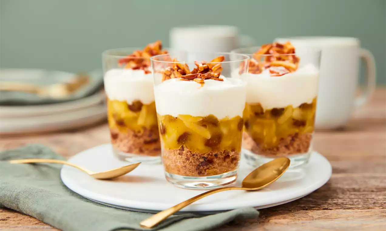 Schichtdessert im Glas mit knusprigem Keksboden, goldgelber Apfelkomponente, weißer Cremehaube und karamellisiertem Topping auf einem Teller