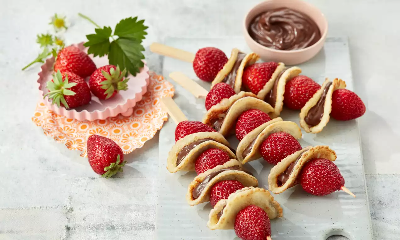 Auf Holzspießen gereihte rote Erdbeeren und goldbraune Püfferchen mit Schokoladencreme, arrangiert auf hellem Untergrund mit Schälchen und Dekoration
