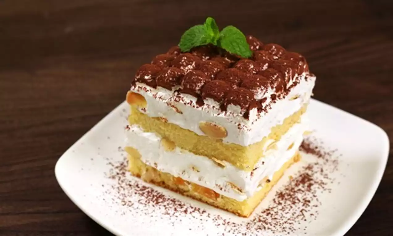 Square tiramisu cake with layers of golden sponge and cream, topped with cocoa powder and a mint sprig on a white plate