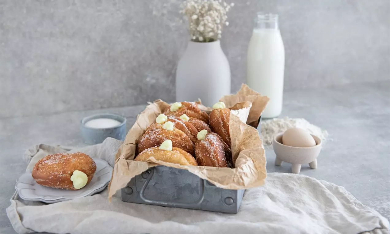 Zlatavé kulaté bombolini poprášené cukrem v kovové nádobě s hnědým papírem, zdobené světlým krémem na neutrálním textilním podkladu