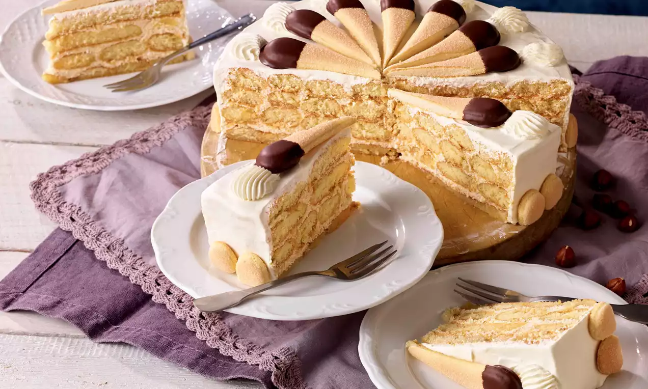 Auf einem weißen Teller liegt ein Stück Malakofftorte mit schichtweise heller Creme und Biskuit, dekoriert mit Schokolade und Biskuitstäbchen