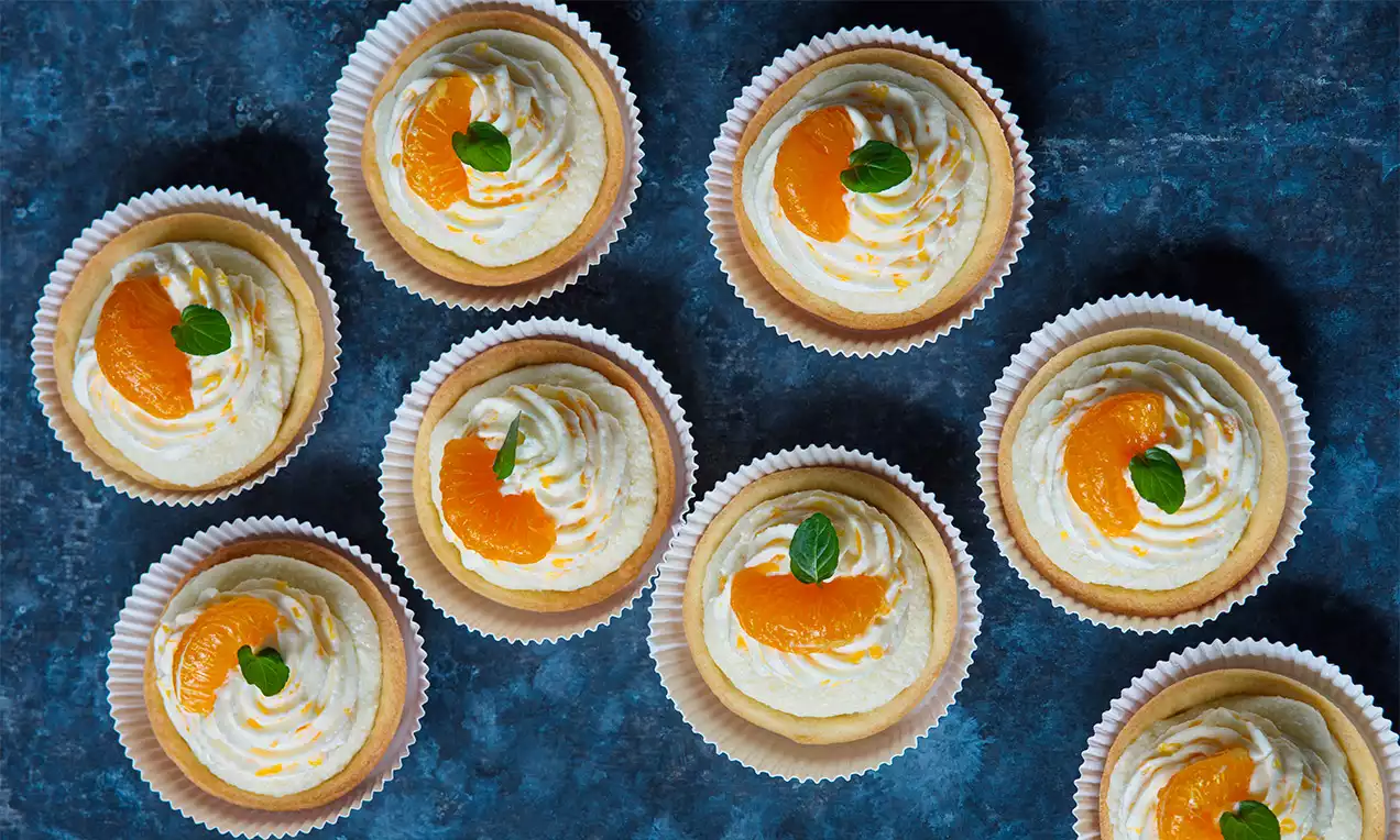 Krehká svetlohnedá tartaletka v papierovom košíčku, s krémovou bielou náplňou, zdobená plátkom mandarínky a lístkom mäty na tmavomodrom podklade