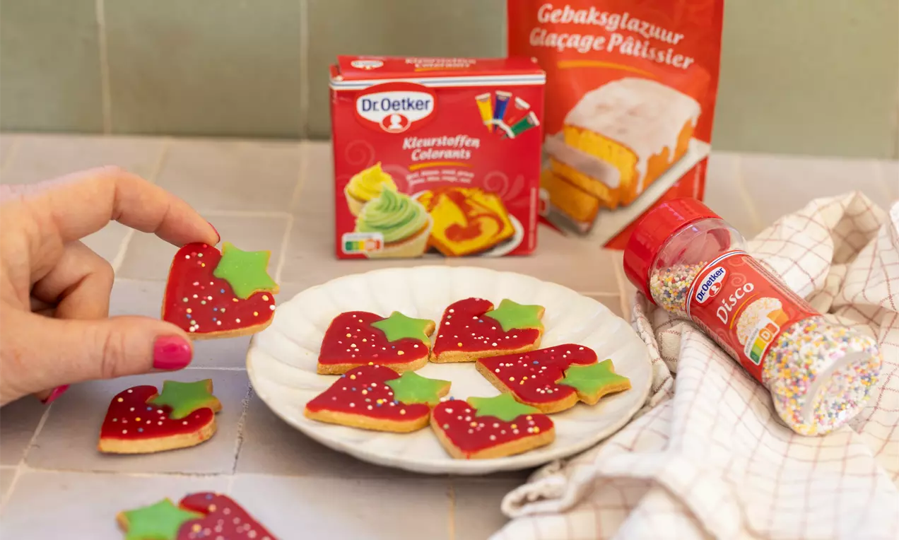 Biscuits en forme de fraises rouges décorées de glaçage et de feuilles vertes, posés sur une assiette blanche en céramique
