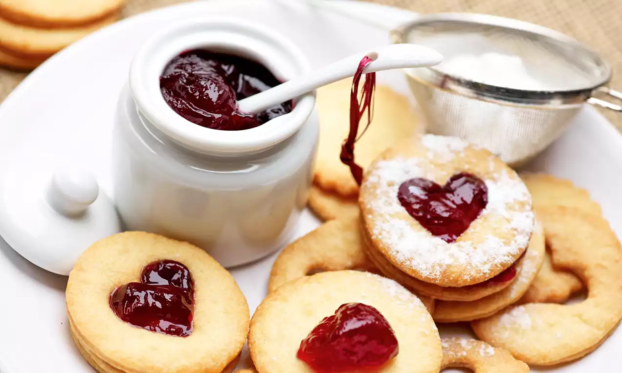Kruche, złociste ciasteczka w kształcie serc z wiśniową galaretką na białym talerzu, obok ceramiczny słoiczek z galaretką i sitko z cukrem pudrem