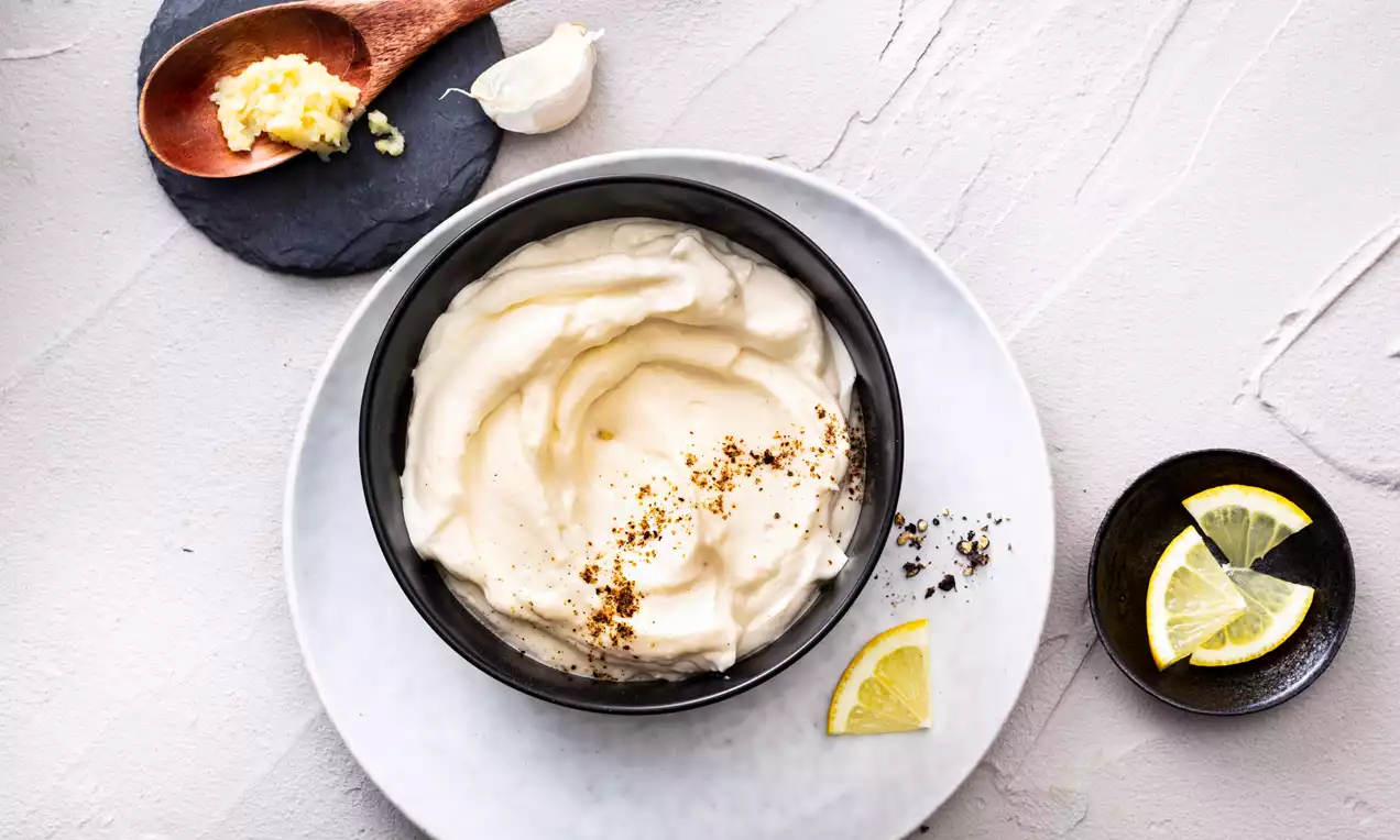 Crème d'ail onctueuse couleur ivoire dans un bol noir, saupoudrée d'épices, avec quartiers de citron et ail sur une assiette blanche