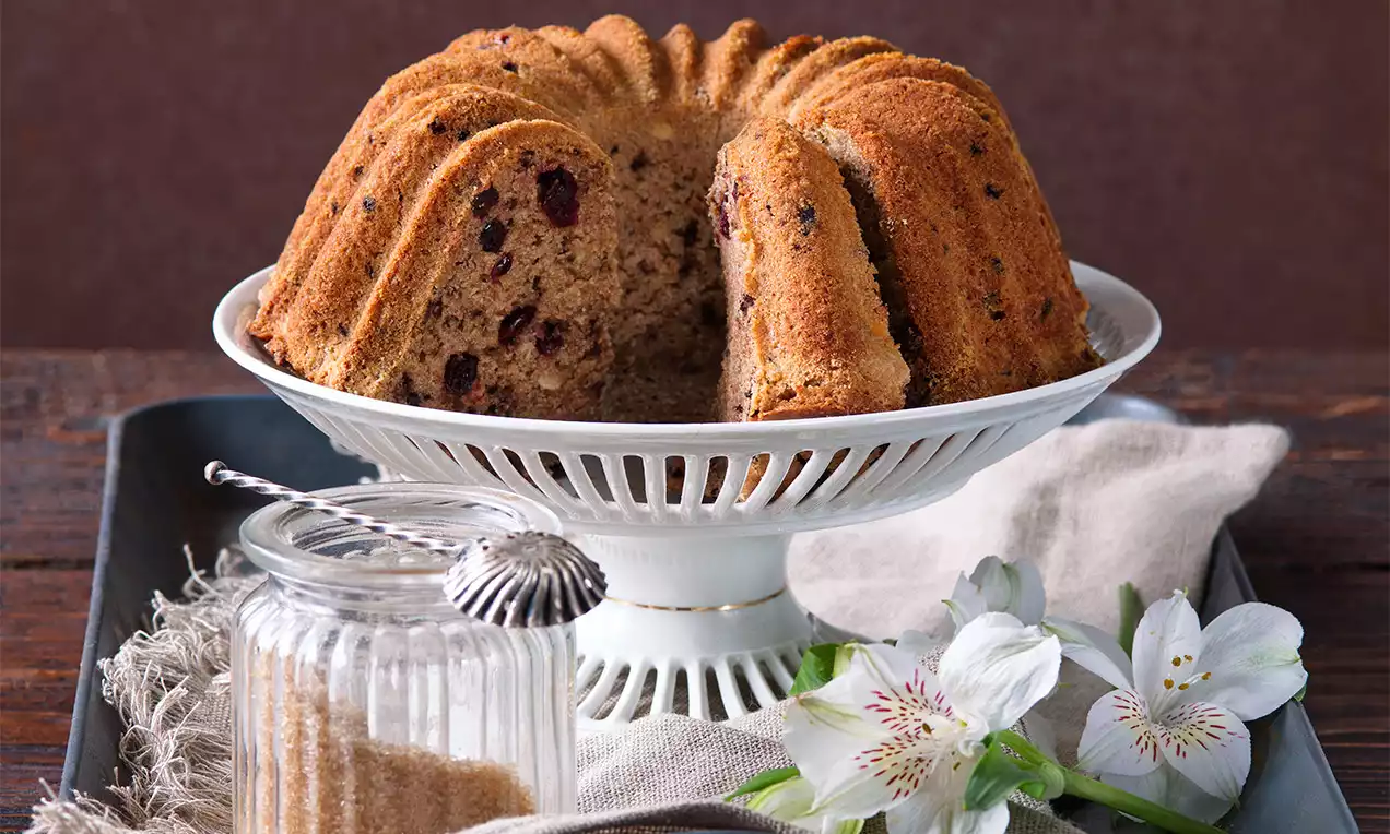 Banánová bábovka zlatohnedej farby s jemne vláčnou textúrou, plátky odhalené, na bielom keramickom stojane vedľa sklenenej dózy a kvetov