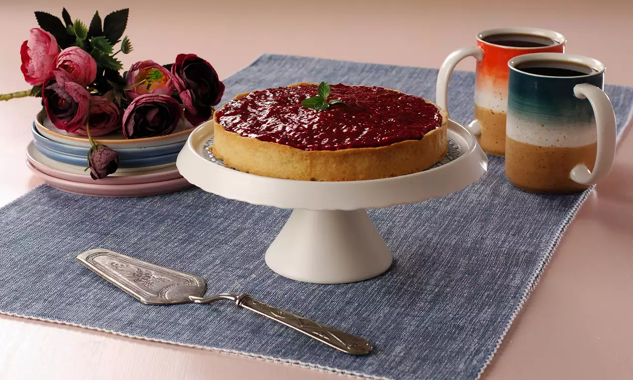 Yuvarlak beyaz sunum tabağında yer alan altın kahverengi hamurlu tart, parlak kırmızı ahududu dolgusu ve üstünde nane yaprağı ile süslenmiş