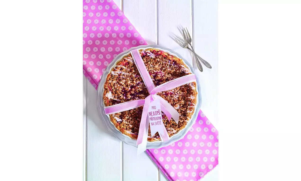 Kulatý švestkový koláč s drobenkou ve skleněné formě, ozdobený růžovou mašlí na růžovém puntíkovaném ubrusu s dvěma vidličkami