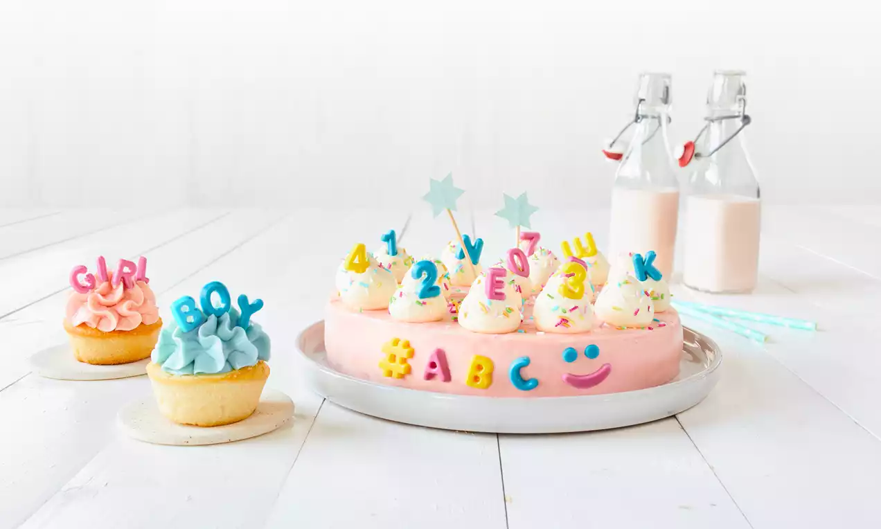Vaaleanpunainen pyöreä kakku valkoisilla kermapursotuksilla, värikkäitä kirjaimia ja numeroita sekä tähtikoristeita vaalealla lautasella