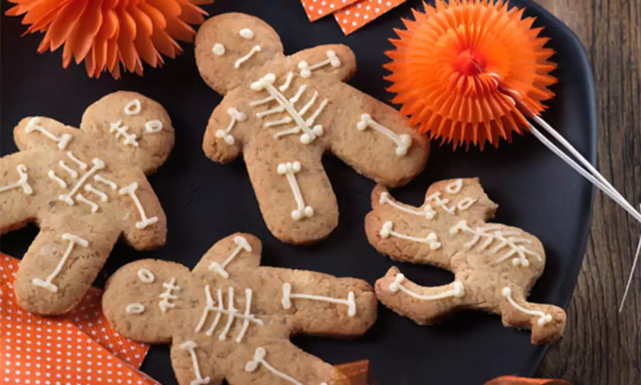 Galletas marrón dorado en forma de figuras humanas y un gato, decoradas con glaseado blanco simulando esqueletos sobre un plato negro