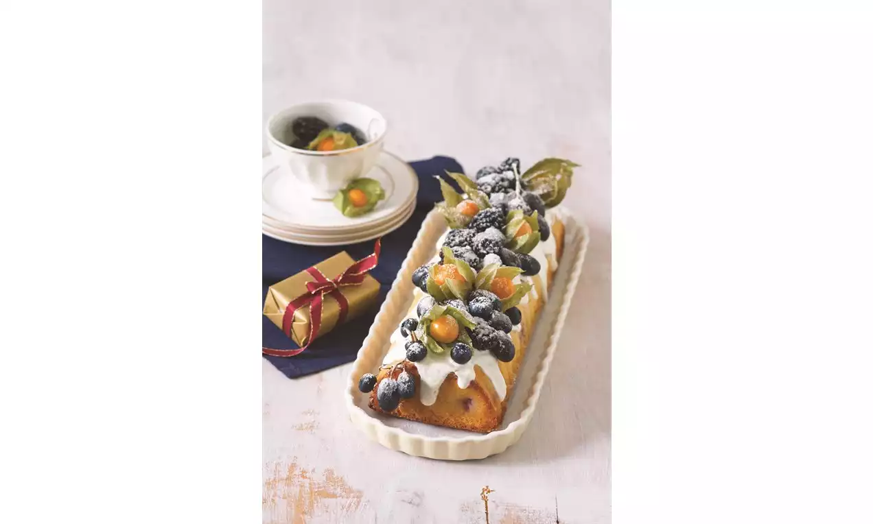 Obdélníkový hřbet zdobený bílou jogurtovou polevou, čerstvým hroznovým vínem, borůvkami, physalis a posypaný moučkovým cukrem na světlém podkladu