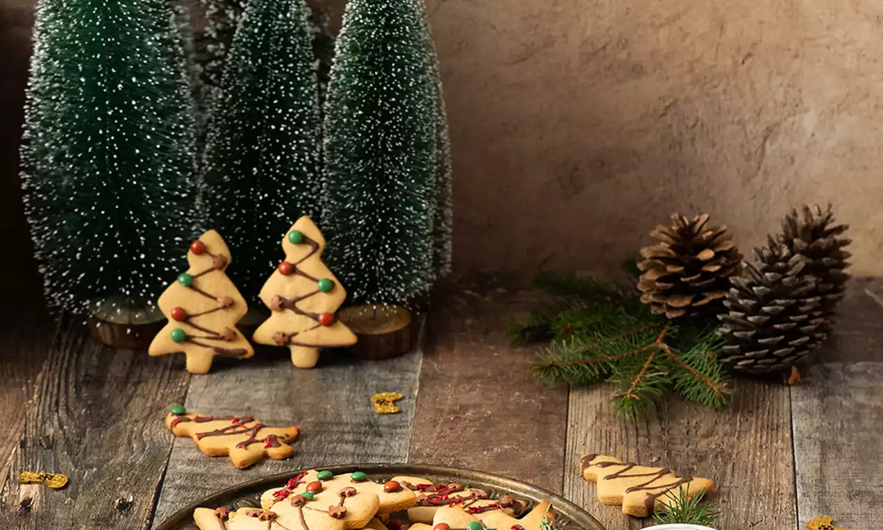 Keksići svijetlosmeđe boje u obliku božićnih drvceta, ukrašeni šarenim glazurnim detaljima na rustikalnom drvenom stolu s borovima i češerima