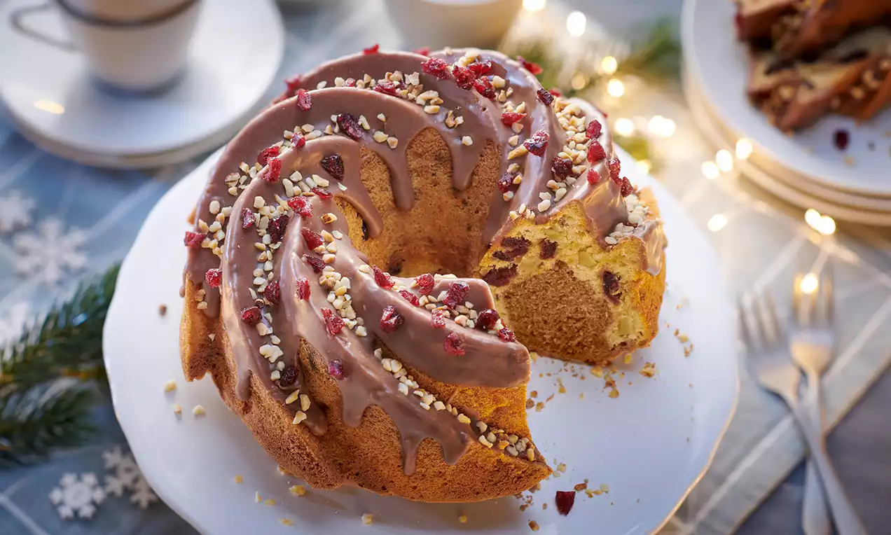 Apfel-Zimt-Gugelhupf mit goldgelber Krume, braunem Zuckerguss, roten Beeren und gehackten Nüssen auf weißer Kuchenplatte mit Tannenzweig