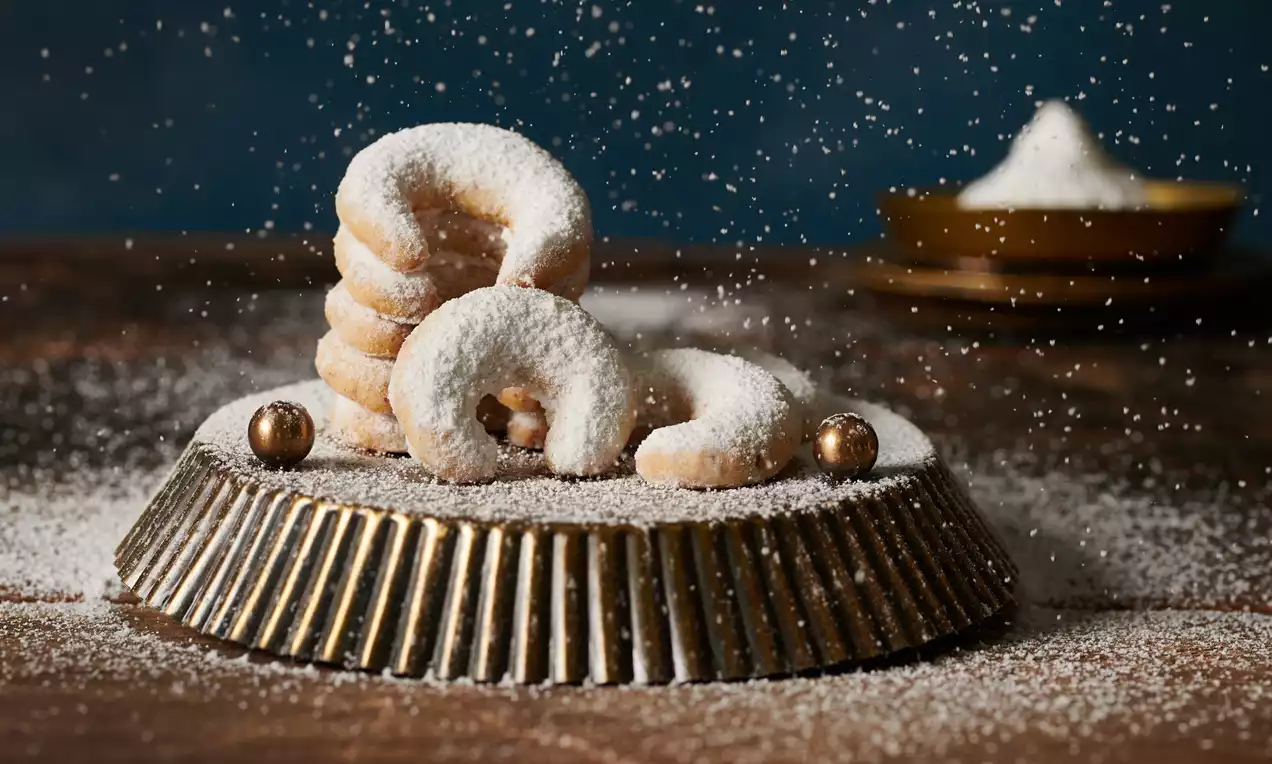 Vanilkové rožky svetlej farby, krehké, zahnuté do jemného oblúka, zasypané práškovým cukrom, naaranžované na kovovom podnose na drevenom stole