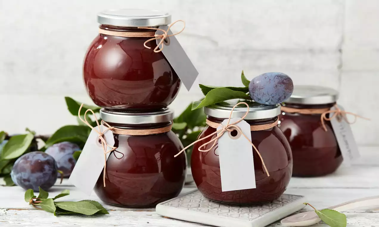 Rundliche Gläser mit glänzend dunkelrotem Zwetschkenmus, beschriftete Anhänger aus Papier, dekoriert mit frischen Zwetschken und grünen Blättern