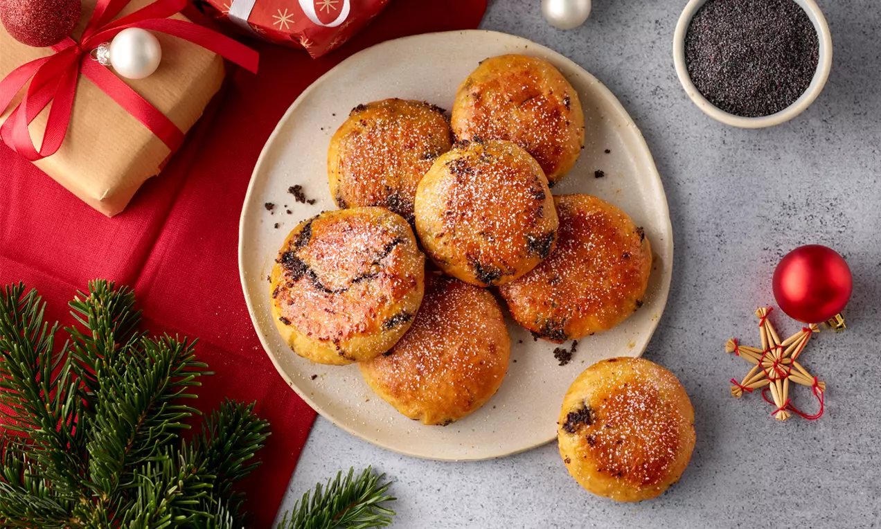 Goldbraune, runde Mohnzelten mit glatter Oberfläche und sichtbarem dunklem Mohn, leicht mit Zucker bestäubt auf einem Teller mit Weihnachtsdekoration
