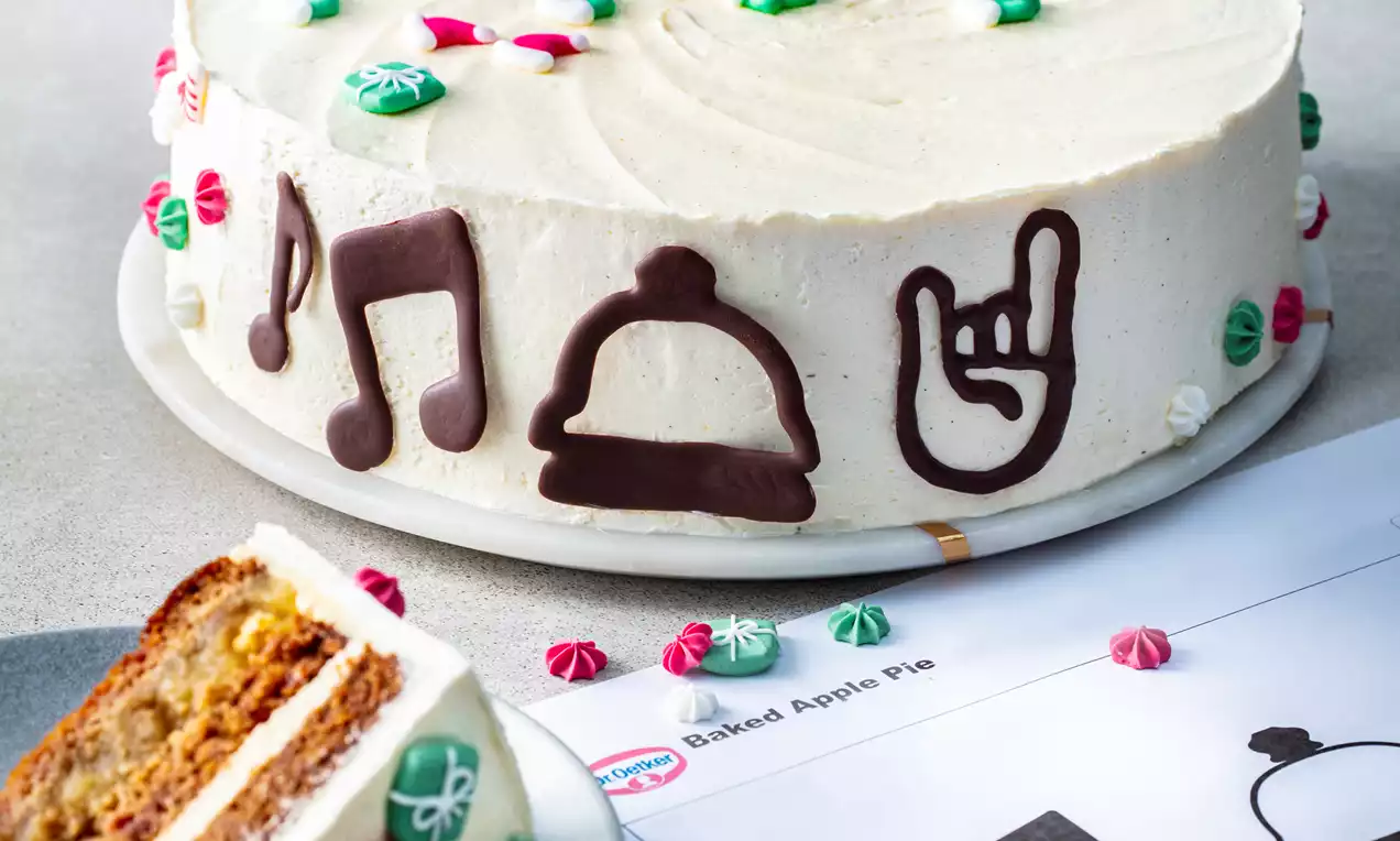 Round layered apple cake covered in smooth white frosting with dark chocolate music and holiday-themed motifs, topped with small red and green decorative dollops