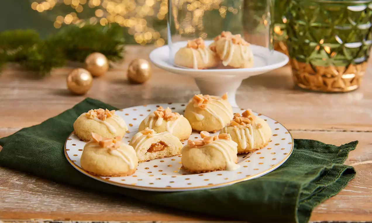 Gefüllte Karamellkekse mit goldbraunen Krümeln und heller Glasur auf einem weißen Teller mit goldenen Punkten auf grünem Stoff drapiert