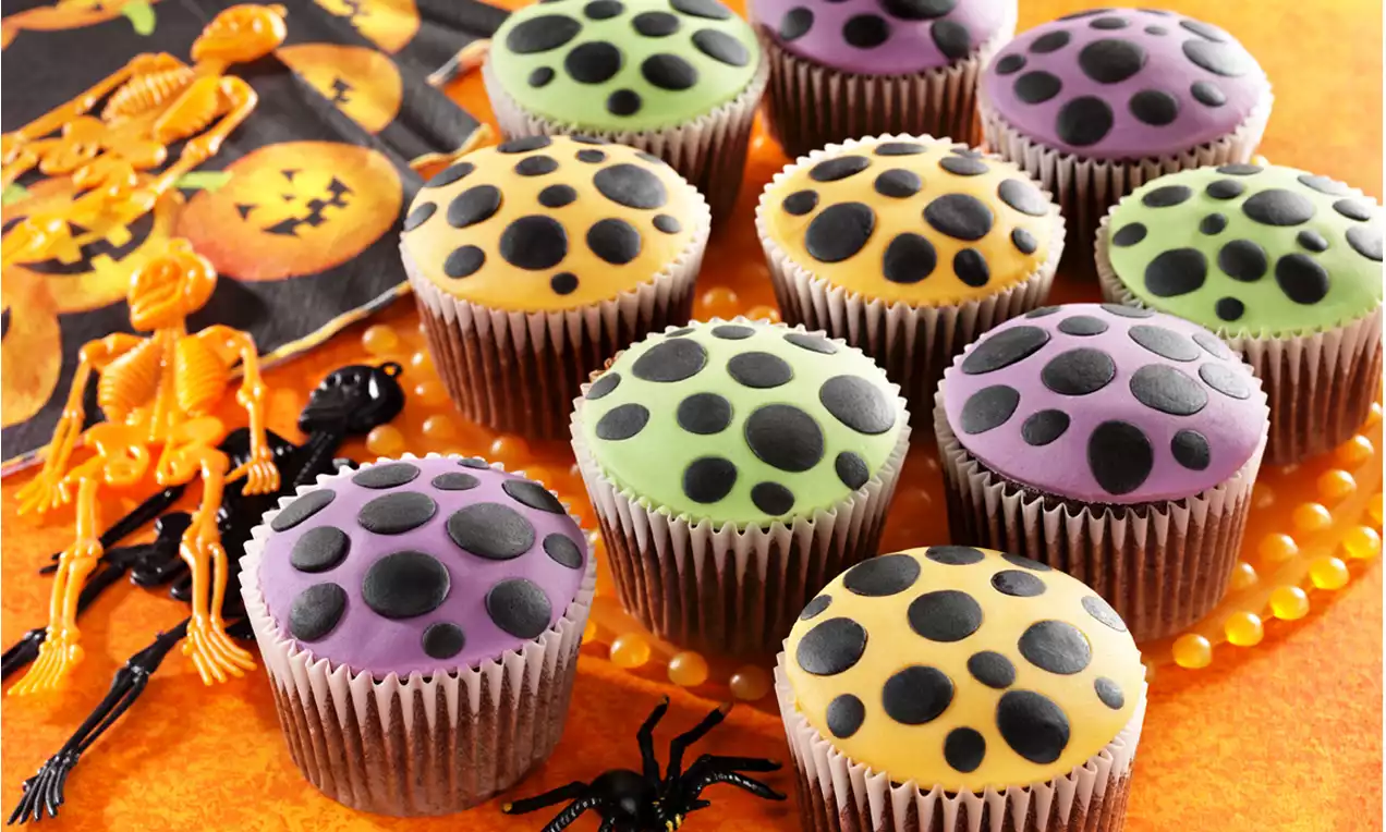 Bunte Cupcakes mit schwarzem Punktmuster in Papierförmchen auf orangefarbenem Hintergrund, dekoriert mit Spinnen und Halloween-Figuren