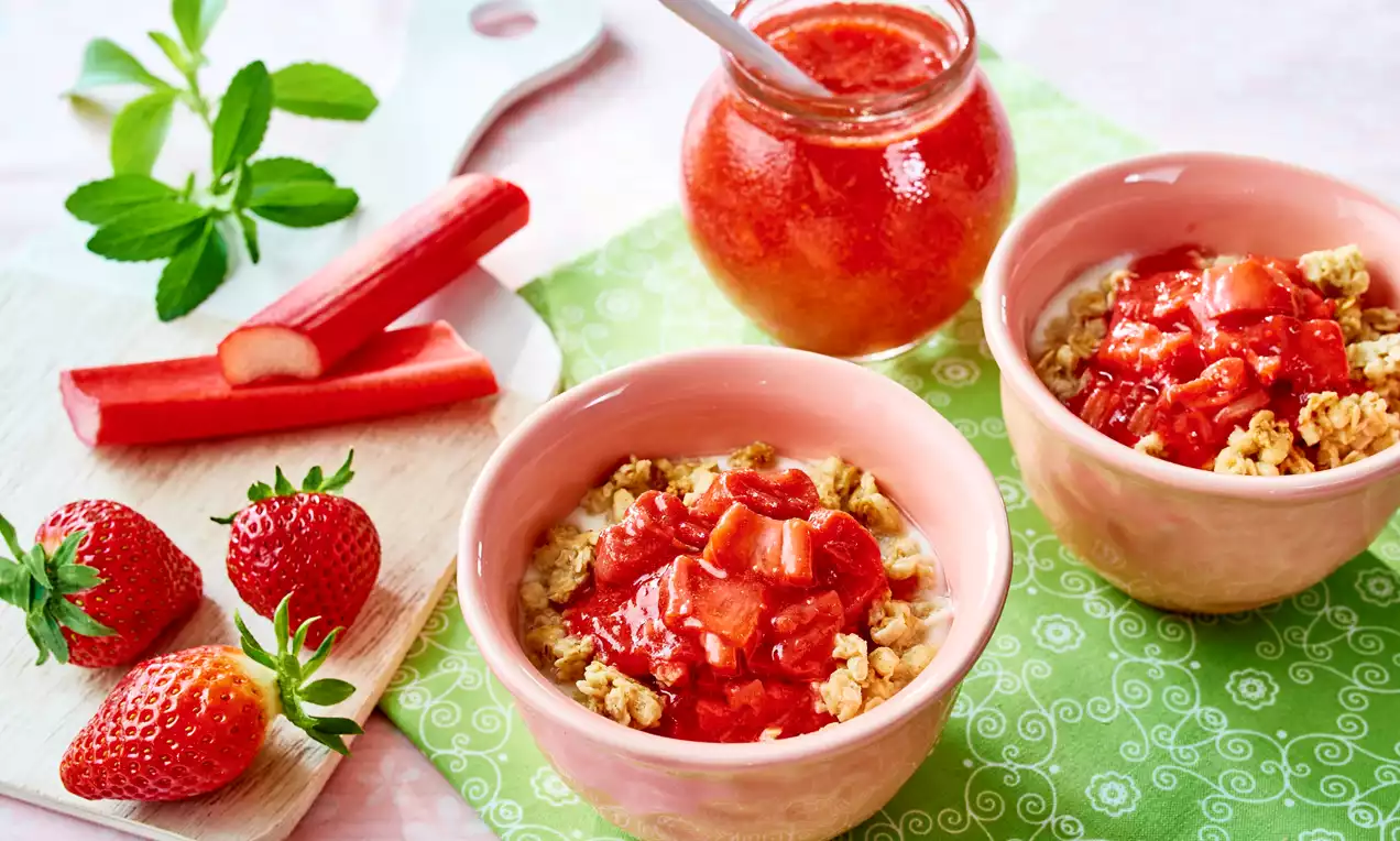 Dżem truskawkowy z rabarbarem w szklanym słoiku oraz w różowych miseczkach z musli, otoczony świeżymi truskawkami i łodygami rabarbaru