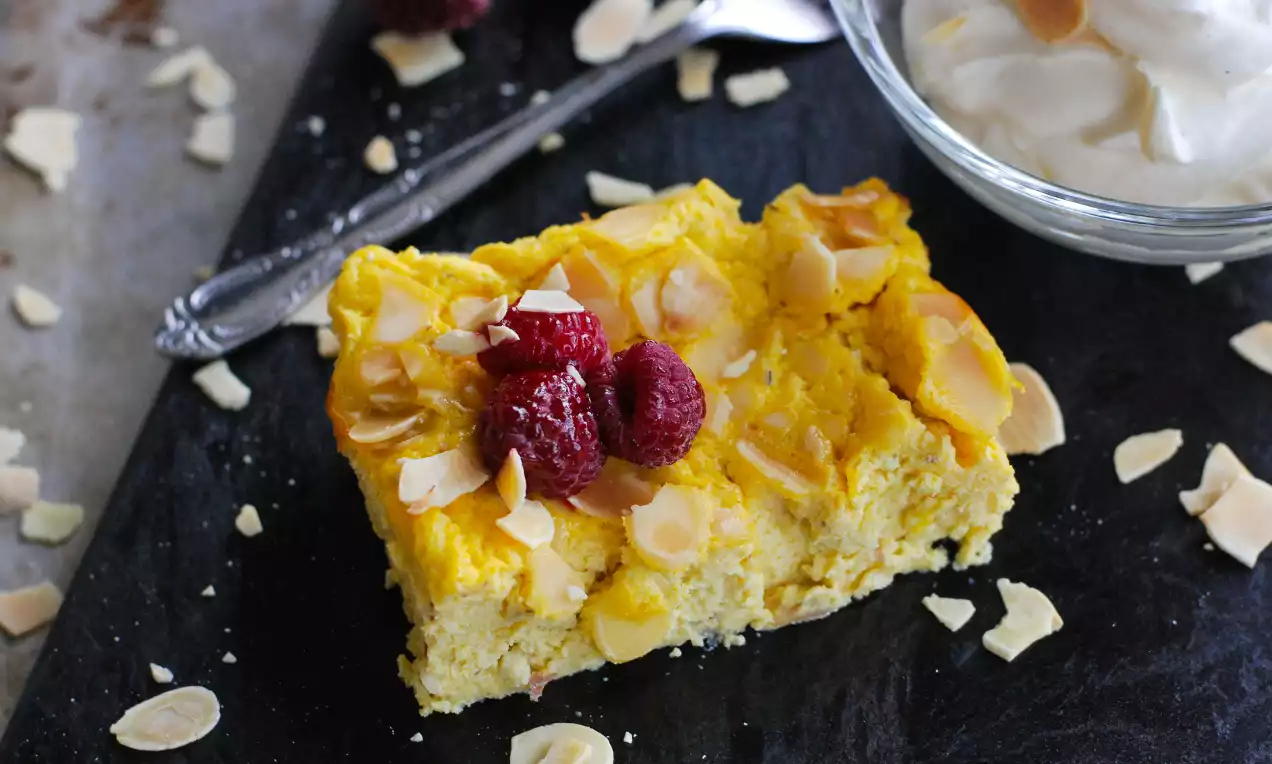 Rektangulär saffransgul ostkaka med mandelspån och hallon på svart skiva, bredvid skål med vispgrädde och en dekorerad sked
