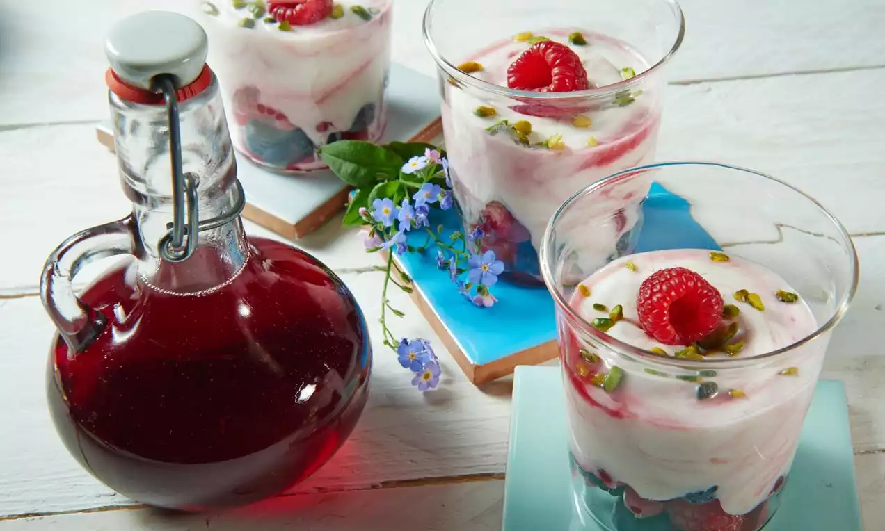 Sticlă rotunjită cu sirop de zmeură rubiniu intens, alături de pahare transparente cu straturi de cremă albă și zmeură roșie viu colorată