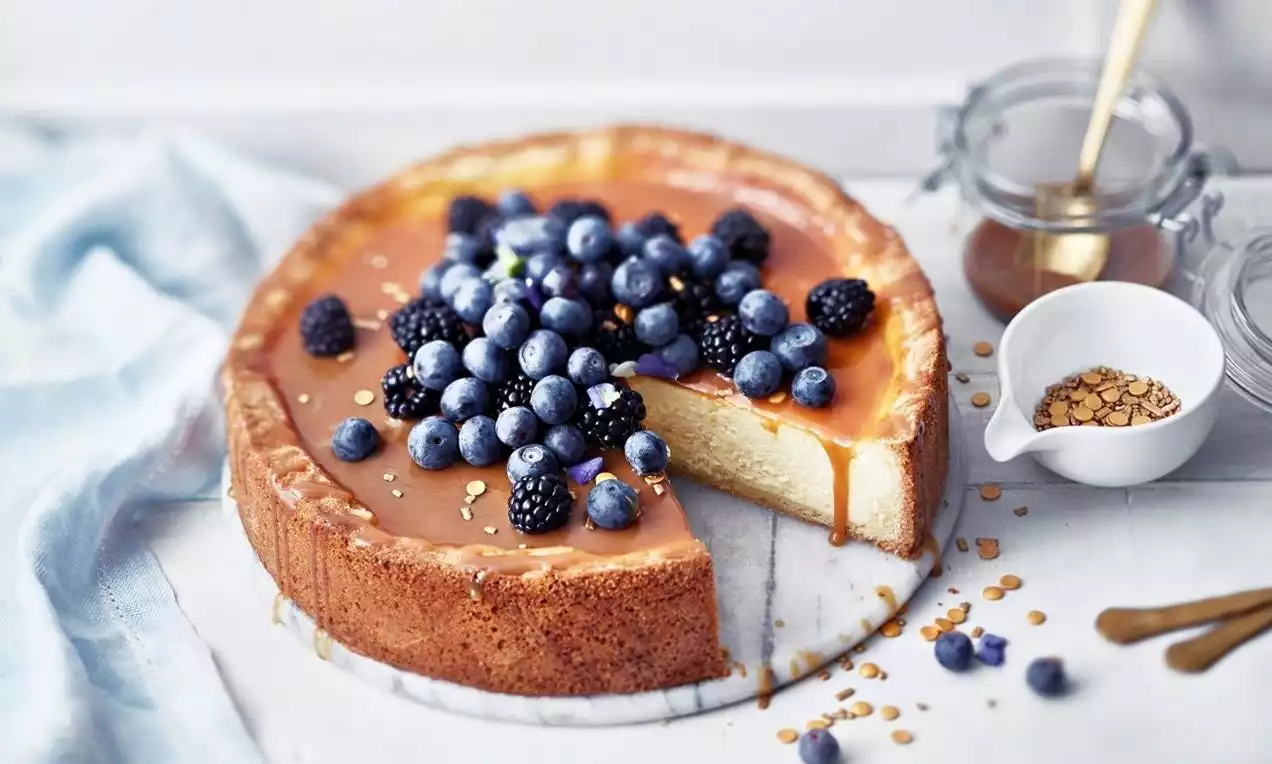 Bakad cheesecake med gyllene yta och kolasås, toppad med blåbär och björnbär, placerad på vit marmorskiva med blå servett