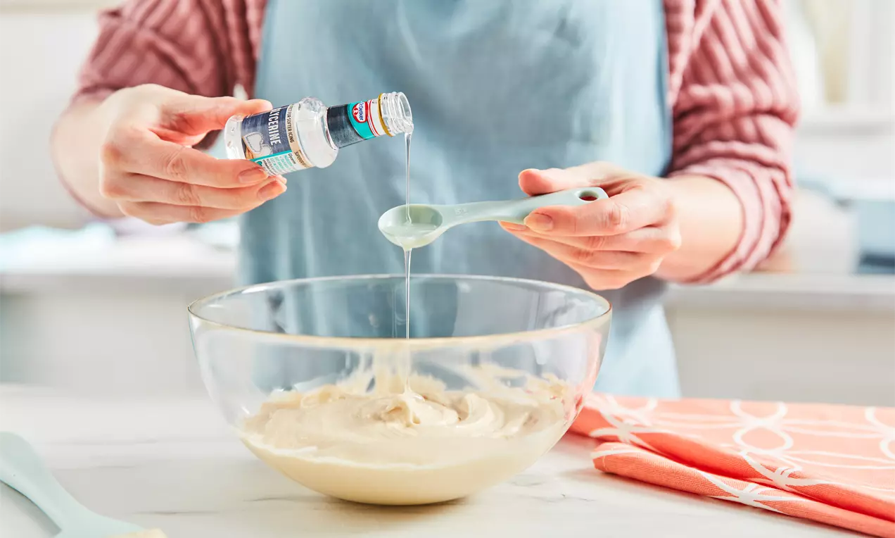 A person in a pale blue apron pouring clear glycerine from a small bottle into a spoon over a glass bowl filled with beige batter