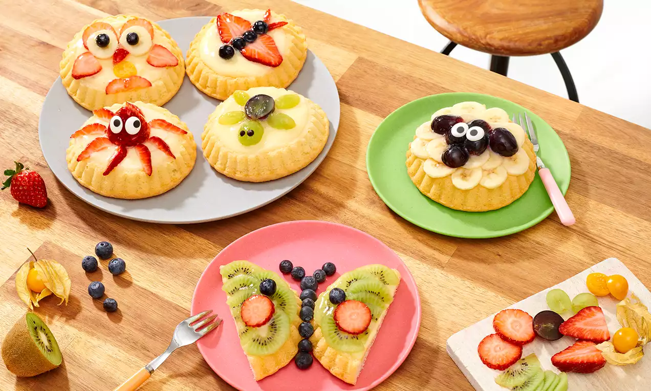 Puddingtörtchen mit bunter Obstdekoration in Tiermotiven, auf Tellern arrangiert und mit frischem Obst auf einem Holztisch drapiert