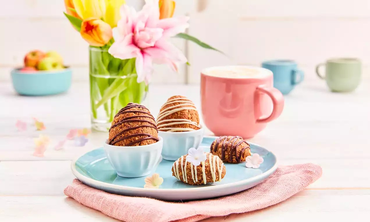 Granola-Eier in kräftigem Braun mit hellen Glasurstreifen, arrangiert auf einem hellblauen Teller mit rosa Serviette und Frühlingsblumen im Hintergrund