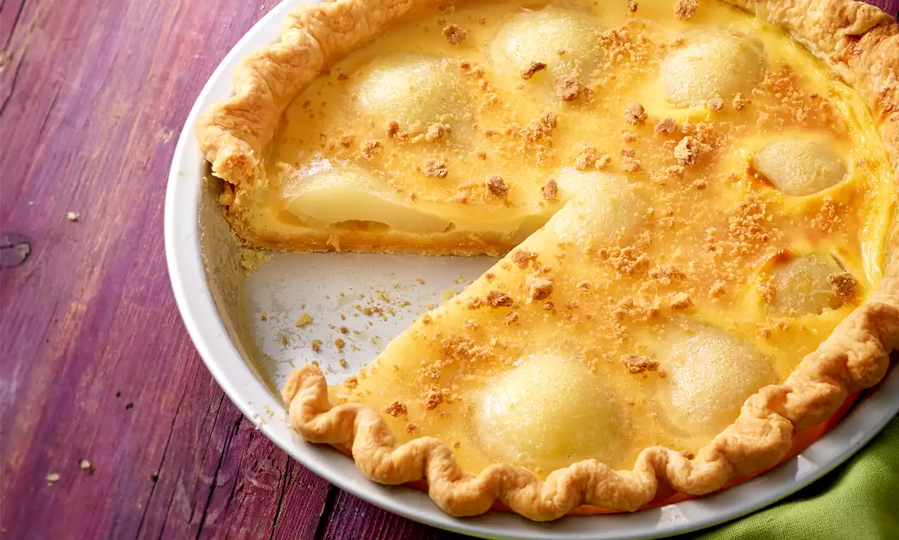 Eine goldgelbe Joghurt-Tarte mit weichen Birnenhälften und bröseligem Belag in einer weißen Backform auf einem violetten Holztisch