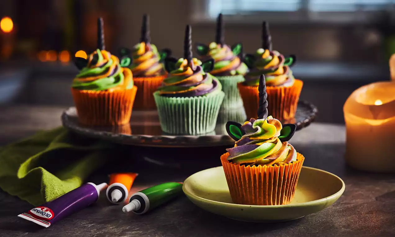 Halloween-themed cupcakes in orange and green cases topped with swirls of purple, green, and orange frosting, black unicorn horns, and ears
