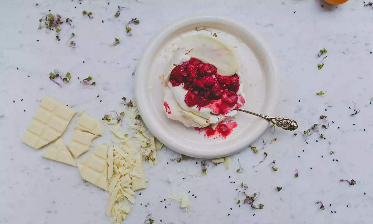 Un dessert bianco cremoso con cioccolato bianco a scaglie, ribes rossi succosi e un cucchiaio d’argento su un piatto bianco marmorizzato