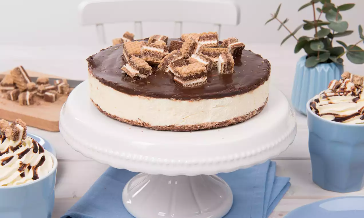 Knoppers-Torte mit brauner Schokoglasur und dekorativen Knoppers-Stückchen auf weißer Tortenplatte, umgeben von blauen Tassen und Tellern